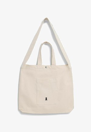 Beige canvas tas met voorvak, korte handgrepen, lange schouderriem en klein zwart logo in het midden van het vak.