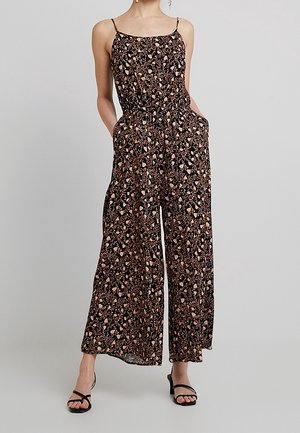 Frau trägt schwarzen ärmellosen Jumpsuit mit pink-beigem Blumenmuster, weitem Bein und Taschen, kombiniert mit schwarzen Riemchenabsatzsandalen.