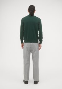 Groen gebreide trui met lange mouwen en geribbelde zoom, gecombineerd met grijze plooibroek, gedragen met zwarte schoenen. Van achter bekeken.