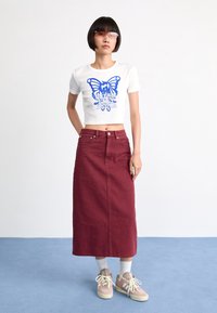 Vit croppad t-shirt med blå fjärilsgrafik, kombinerad med en vinröd jeansmaxikjol. Modellen bär cremefärgade sneakers och vita strumpor.