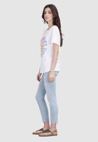 T-shirt blanc à manches courtes avec un graphique circulaire coloré, associé à un jean slim bleu clair et des sandales métalliques.