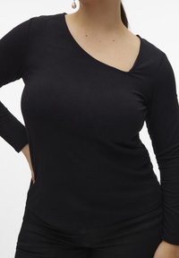 Top nero a maniche lunghe a costine con scollatura asimmetrica, texture liscia e design aderente. Indossato con pantaloni neri e orecchini a perla minimalisti.