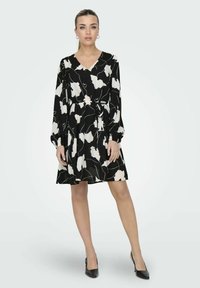 Robe noire avec un motif floral blanc, décolleté en V, manches bouffantes longues, ceinture à nouer à la taille et jupe évasée. portée avec des chaussures à talons hauts noires.