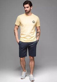 T-shirt in cotone giallo con logo rotondo, abbinato a pantaloni corti blu navy e sneaker grigio chiaro. Maniche corte e vestibilità comoda.