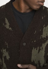 Brauner gestrickter Cardigan mit grünem Camouflage-Muster, verfügt über eine strukturierte Oberfläche, einen tiefen V-Ausschnitt und einen einzelnen Knopfverschluss.