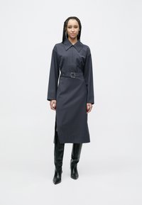 Námornícka košeľová šaty s dlhými rukávmi, špicatým golierom, opaskom v páse, bočnými rozparkami a modelkou v čiernych čižmách po kolená. Hladký povrch.