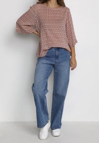 Chemisier rouge et blanc à motif avec des manches 3/4, associé à un jean bleu taille haute et des baskets blanches, le tout sur un fond neutre.