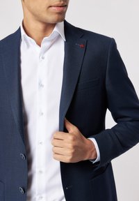Marineblauer strukturiertes Sakko mit zwei Knöpfen, getragen über einem weißen Hemd. Enthält ein kleines rotes Logo am Revers und sichtbare Details an den Manschetten.