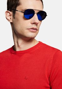 Okrągłe okulary przeciwsłoneczne z niebieskimi soczewkami i cienkimi czarnymi metalowymi oprawkami, w połączeniu z czerwonym swetrem z okrągłym dekoltem z małym logo na piersi.