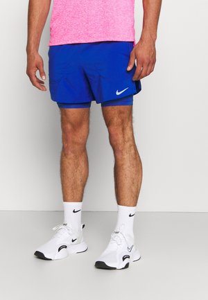 Mężczyzna w jasnoróżowej koszuli, niebieskich krótkich spodenkach do biegania Nike, białych skarpetkach Nike i białych butach do biegania Nike, stojący na jasnoszarej podłodze.