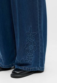 Jean bleu foncé à jambes larges avec des motifs floraux brodés près de l'ourlet, associé à des chaussures noires à bouts carrés sur fond blanc.