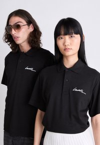 Dos modelos vistiendo camisetas polo negras con un logo en blanco, uno con gafas de sol y cabello rizado, y el otro con cabello lacio negro y una falda blanca.