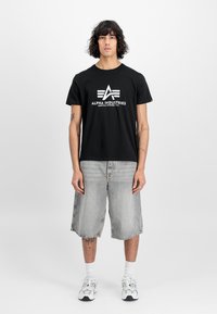 Sort bomuld t-shirt med hvidt "ALPHA INDUSTRIES" logo, parret med grå denimshorts og hvide ankelstrømper, båret med grå sneakers.