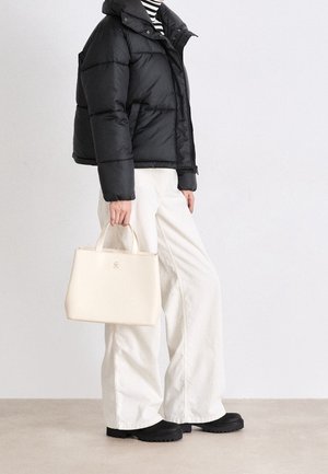 Personne portant une doudoune noire, des bottes noires, un pantalon blanc à jambes larges, une chemise rayée, tenant un sac à main couleur crème sur un fond uni.