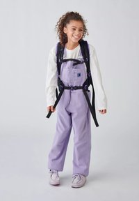 Lila Latzhose mit verstellbaren Trägern, vorderseitigen Taschen und schwarzem Zubehör, kombiniert mit einem weißen Langarmshirt und hellen Sneakers.