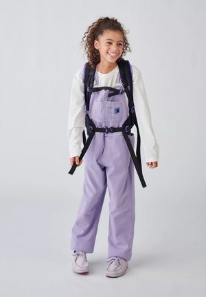 Salopette lilla con spalline regolabili, tasche frontali e accessori neri, abbinata a una camicia bianca a maniche lunghe e sneakers chiare.