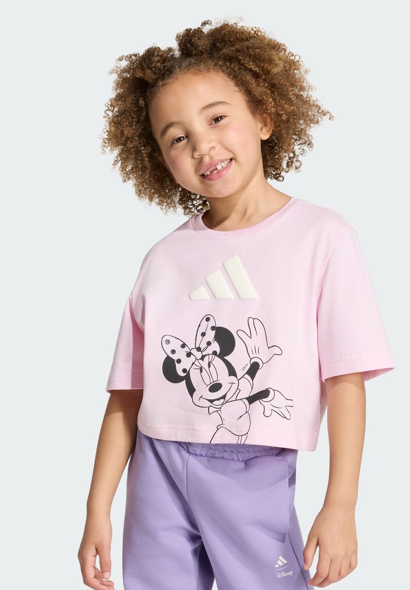Enfant souriant aux cheveux bouclés portant un t-shirt court rose avec Minnie Mouse et un short violet Adidas.