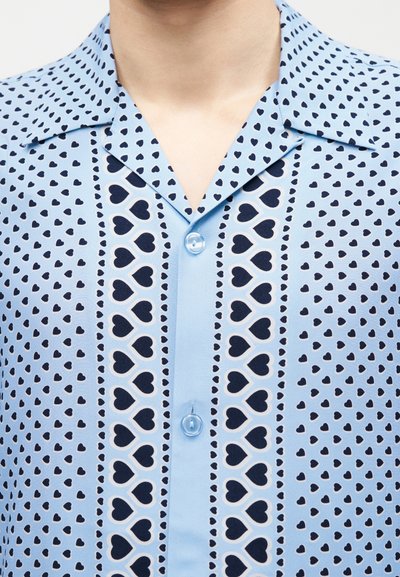 Φωτεινό γαλάζιο πουκάμισο με γιακά, με κάθετες σειρές από μεγάλες και μικρές σκοτεινές μπλε καρδιές και κουμπιά σε αντίστοιχο χρώμα.