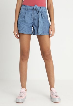 Kvinde iført blå stribede shorts med høj talje og bælte, rød top og lyserøde sneakers står mod en hvid baggrund.