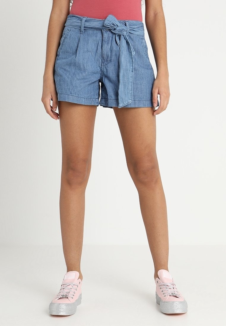 Femme portant un short bleu à rayures taille haute avec ceinture, un haut rouge et des baskets roses, debout sur un fond blanc.