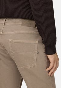 Pantalones de algodón beige con un diseño texturizado y bolsillos traseros, presentando una etiqueta de cuero marrón estampada con "B BOGGI MILANO."