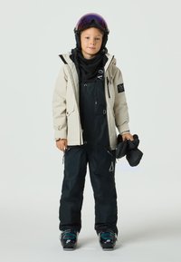 Barn som bär en ljusbeige vattentät jacka över en svart skidoverall med justerbara remmar, håller svarta handskar. Skidglasögon vilar på huvudet.