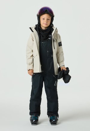 Kind trägt eine hellbeige wasserdichte Jacke über einem schwarzen Schneeanzug mit verstellbaren Trägern und hält schwarze Handschuhe. Skibrille liegt auf dem Kopf.