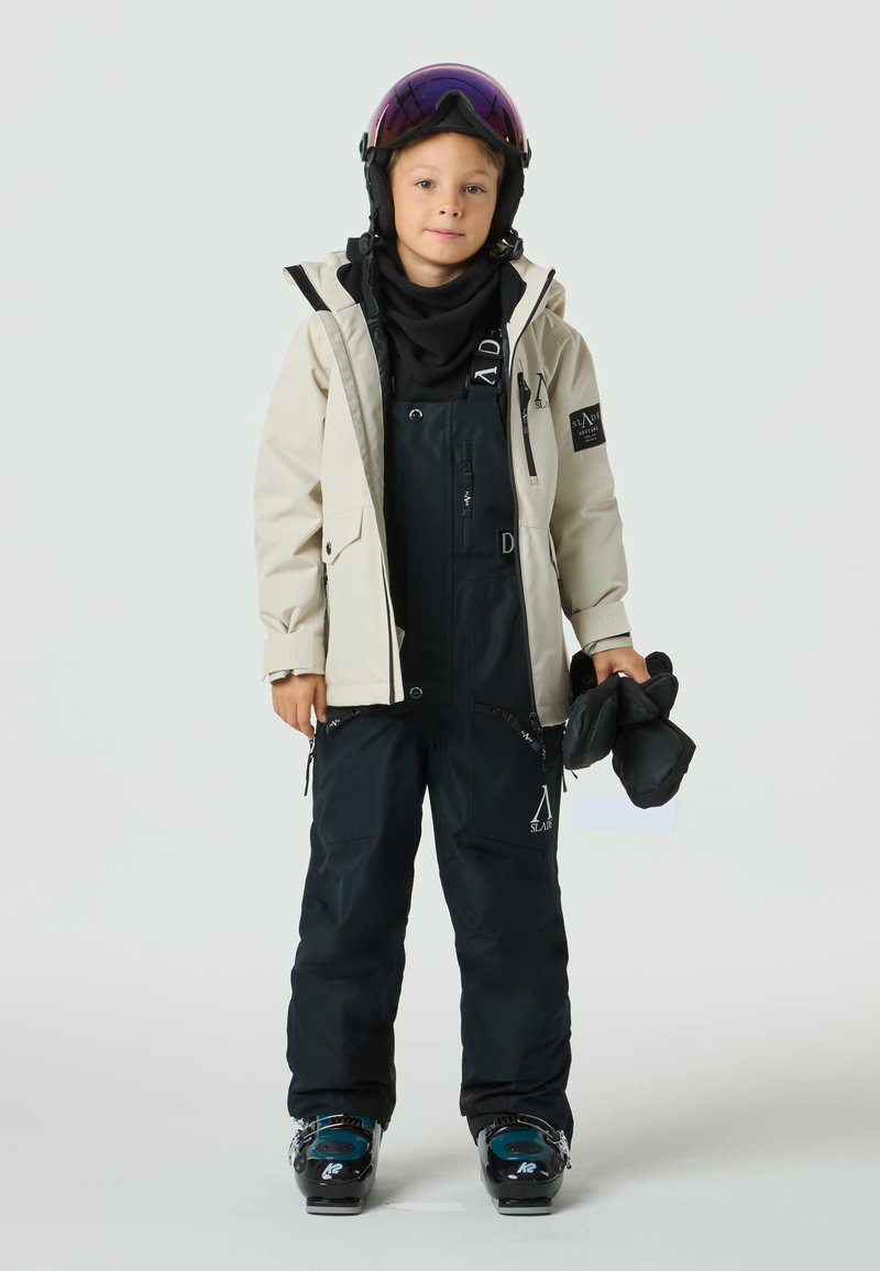 Barn som bär en ljusbeige vattentät jacka över en svart skidoverall med justerbara remmar, håller svarta handskar. Skidglasögon vilar på huvudet.