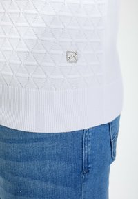 Suéter blanco de punto con un patrón de diamante texturizado, dobladillo acanalado y un pequeño logo plateado. Llevado con jeans de mezclilla azul.