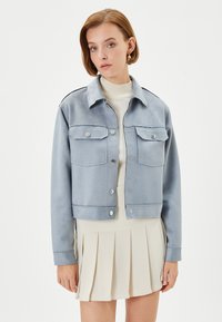 Lichtblauwe jas van zacht materiaal, met voorste drukknoopsluitingen, twee borstzakken en contrasterende donkere schouder- en manchetdetails.