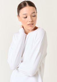 Top bianco a maniche lunghe con vestibilità comoda, caratterizzato da polsini a coste e un ampio scollo. Tessuto liscio con una texture morbida.