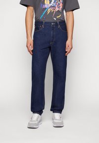 Mörkblå rak ben-jeans med fem fickor, kontrasterande sömmar och metallknappslutning. Bärs med en grafisk grå t-shirt och sneakers.