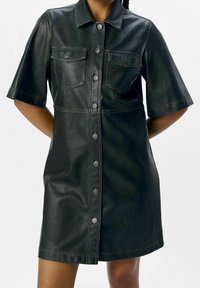Robe chemise en simili cuir noir avec des manches courtes, deux poches poitrine et fermeture à boutons sur le devant. Présente des coutures contrastées.