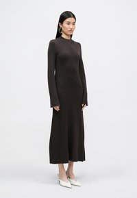 Długa, dopasowana brązowa dzianinowa sukienka z długimi rękawami i bocznymi rozcięciami, zestawiona z białymi czółenkami na wysokim obcasie. Gładka tekstura i minimalistyczny design.