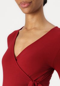 Rotes Wickeloberteil mit tiefem V-Ausschnitt, aus glattem, dehnbarem Stoff. Verfügt über eine seitliche Knoten-Detail, das das figurbetonte Design betont.