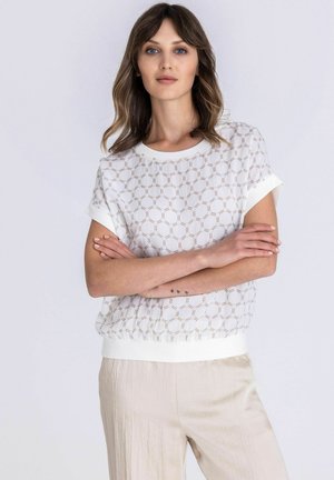 Vrouw met bruin schouderlang haar draagt een wit topje met patroon en korte mouwen en lichtbeige broek, staand met gekruiste armen tegen een witte achtergrond.