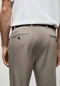Pánské béžové kalhoty s zadními kapsami a černými knoflíkovými detaily. Tkanina je hladká s vypasovaným střihem. Nošeny s bílou košilí s krátkým rukávem.