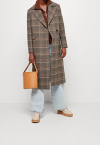 Persoon in een bruin geruite lange jas, lichtblauwe wijde jeans, beige schoenen en een beige emmer tas houdend tegen een witte achtergrond.