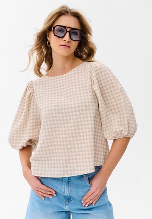 Kobieta o falowanych blond włosach, nosząca duże okulary przeciwsłoneczne w oprawkach w kolorze żółwia, złote kolczyki koła, beżową kratowaną bluzkę z bufkami na rękawach oraz jasnoniebieskie dżinsy.