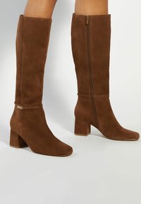Brown suede knee-high boots with a square toe and chunky heel. Features a side zipper and a decorative gold accent near the ankle.