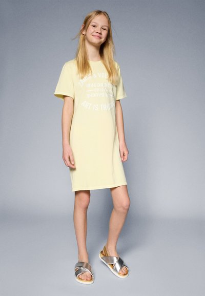 Vestido amarillo de manga corta con texto gráfico en blanco. Combinado con sandalias plateadas de tiras cruzadas y suela de contraste. Textura de tela suave.