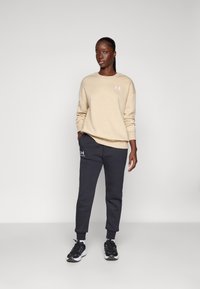 Beige genser med rund hals og logo, kombinert med mørkegrå joggebukser med elastiske mansjetter og logo. Svart joggesko fullfører antrekket.