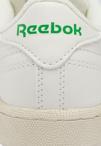 Bijela tenisica s gornjim dijelom od teksturirane kože, zelenim logotipom "Reebok" i kremasto žutom gumom potplatom s malim izdignutim točkicama.