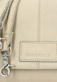 Borsa a tracolla in pelle beige chiaro con superficie testurizzata, dotata di finiture in argento, tracolla rimovibile e etichetta "BURKELY" in rilievo.