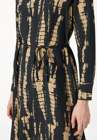 Juoda ir smėlio spalvos raštuota suknelė su ilgomis rankovėmis, susijuosta juosmeniu ir minkšta, tekstūruota medžiaga. Raštas turi nereguliarus vertikalias linijas.