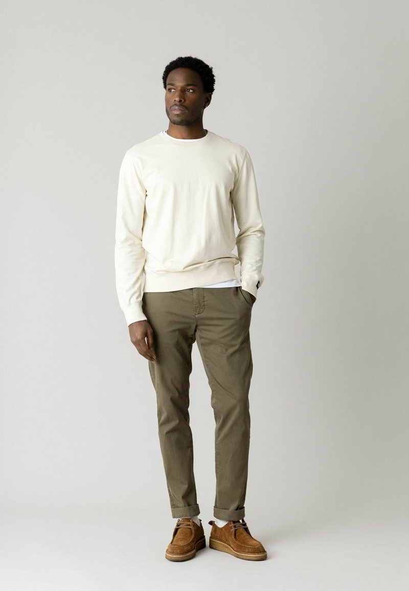 Ung mand med kort krøllet hår står iført en cremefarvet sweater, olivengrønne bukser og brune ruskindssko mod en ensfarvet lys grå baggrund.