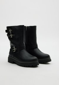 Botas de tobillo de cuero negro con suela gruesa, que cuentan con dos correas ajustables con hebillas y una cremallera lateral. El acabado texturizado añade interés visual.