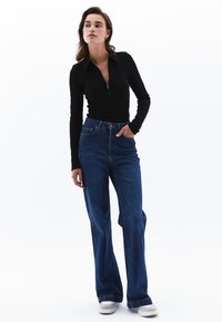 Schwarzes Langarmoberteil mit Reißverschlusskragen; blaue hochgeschnittene Schlagjeans; Model trägt weiße Sneaker, vor einem schlichten weißen Hintergrund.