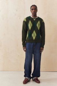 Πλεκτό πουλόβερ σε πράσινο και μαύρο καρό σχέδιο, χαλαρά σκούρα μπλε τζιν και μαρόν παπούτσια. Το μοντέλο στέκεται μπροστά σε ελαφρύ ξύλινο φόντο.