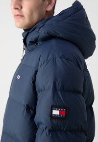Marineblå polstret jakke med hætte, med et logo patch på ærmet. Glat tekstur med quiltet design og snorejusteringer.